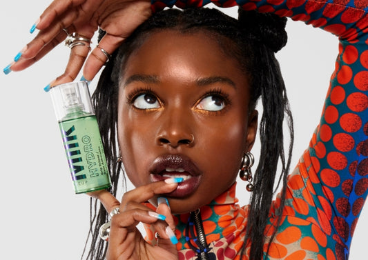 Model in a colorful outfit holds a bottle of Milk Makeup Hydro Grip Primer near her face, against a white background