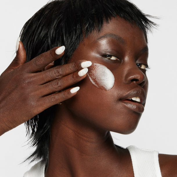 Model applies Milk Makeup Cloud Glow Priming Foam to cheek against a white background